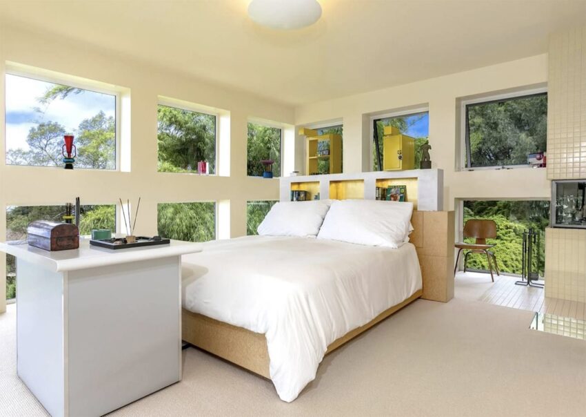 Modern bedroom with large windows, white bedding, a bedside table with decor, and lush greenery visible outside.