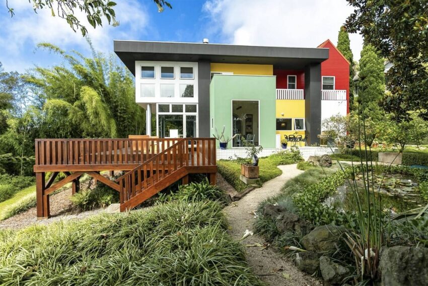Modern multicolored house with large windows and wooden deck, surrounded by lush greenery and a stone path on a sunny day.