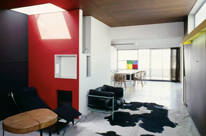 Modern living room with red accent wall, black lounge chairs, cowhide rug, and dining area in the background with colorful art.