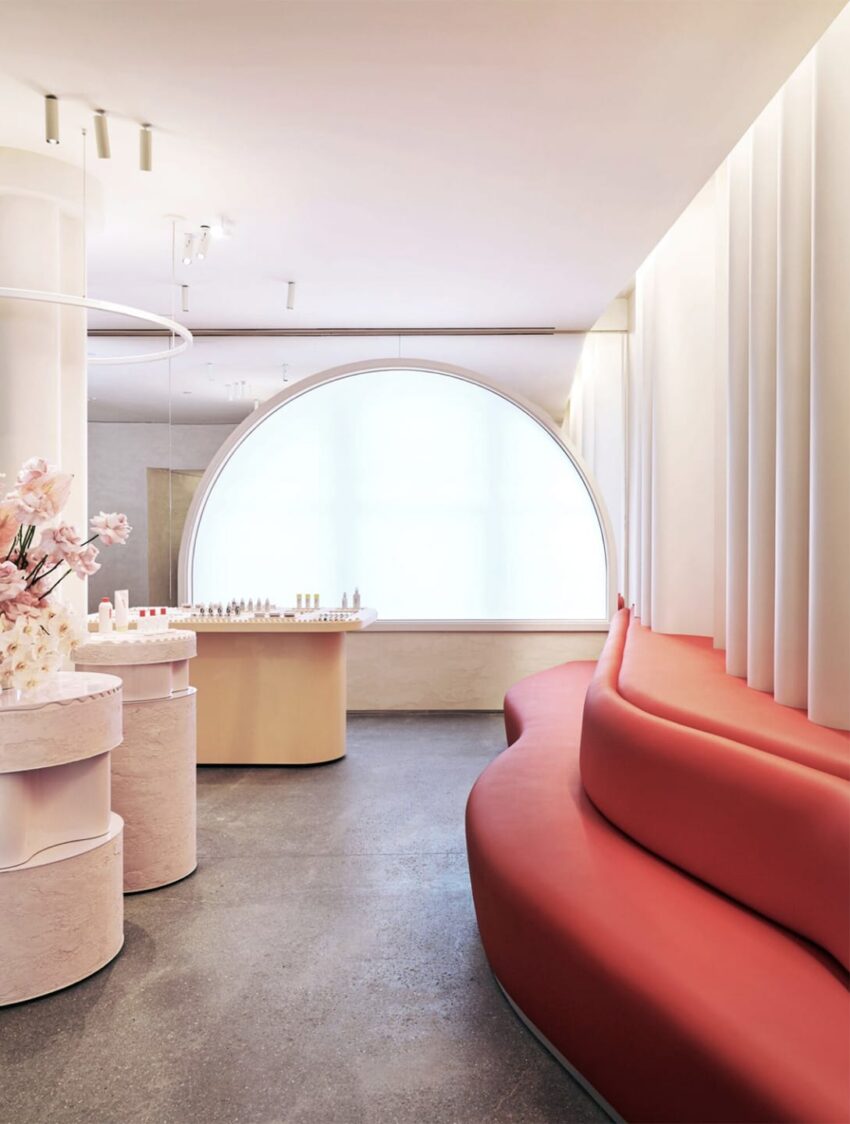 Modern beauty store interior with curved red couch, round display tables, large arched window, and delicate floral decor.