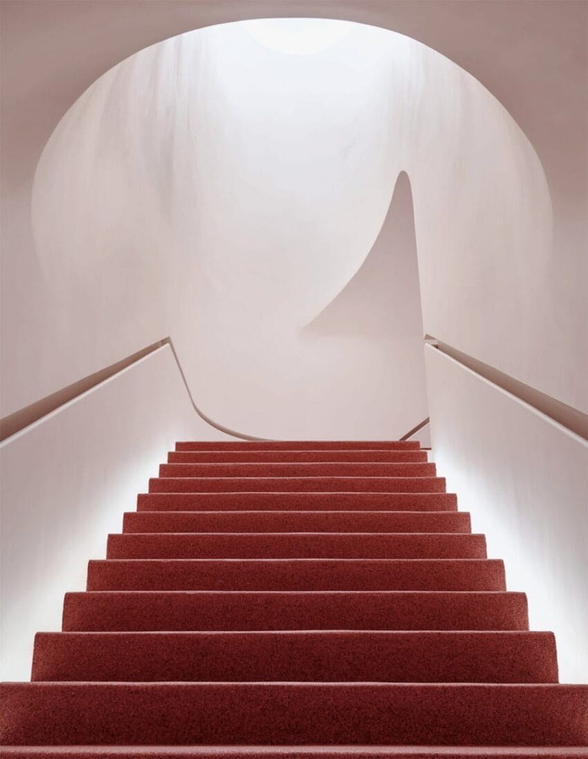 Red carpeted stairs leading upwards in a minimalist setting with curved, smooth beige walls and soft lighting.