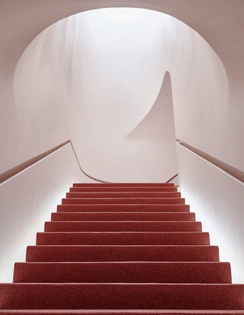 Red carpeted stairs leading upwards in a minimalist setting with curved, smooth beige walls and soft lighting.