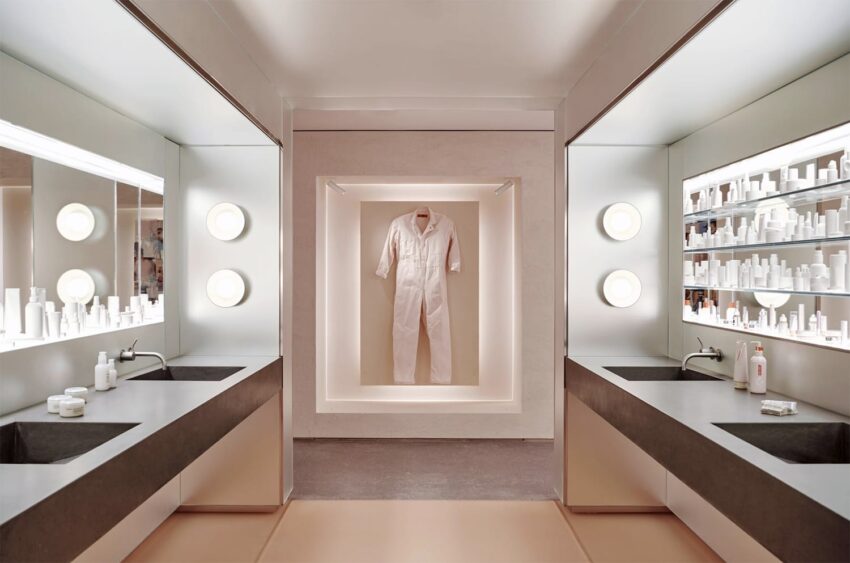 Modern dressing room with a jumpsuit on display, surrounded by illuminated mirrors, sinks, and skincare products.