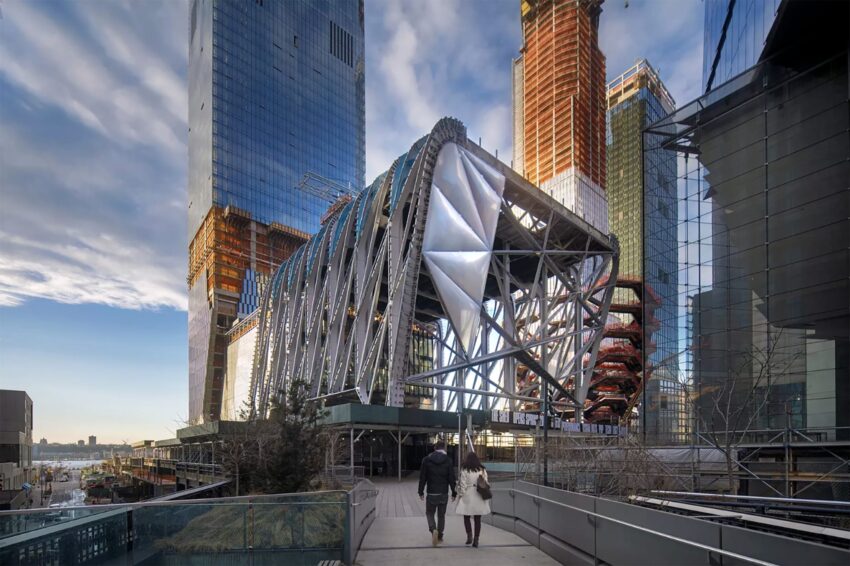 Modern architectural building with metallic exterior, surrounded by skyscrapers, and two people walking towards it.
