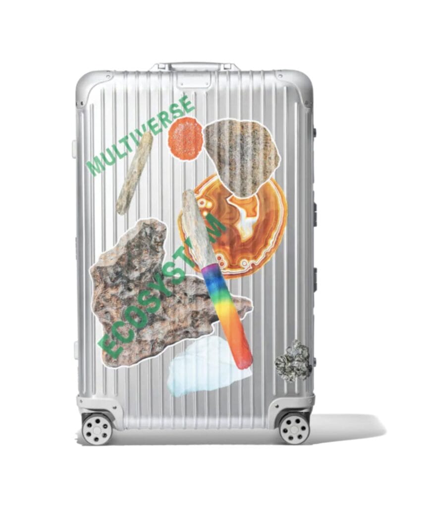 Silver suitcase with colorful sticker designs including rocks, a rainbow, and text reading "Multiverse Ecosystem."
