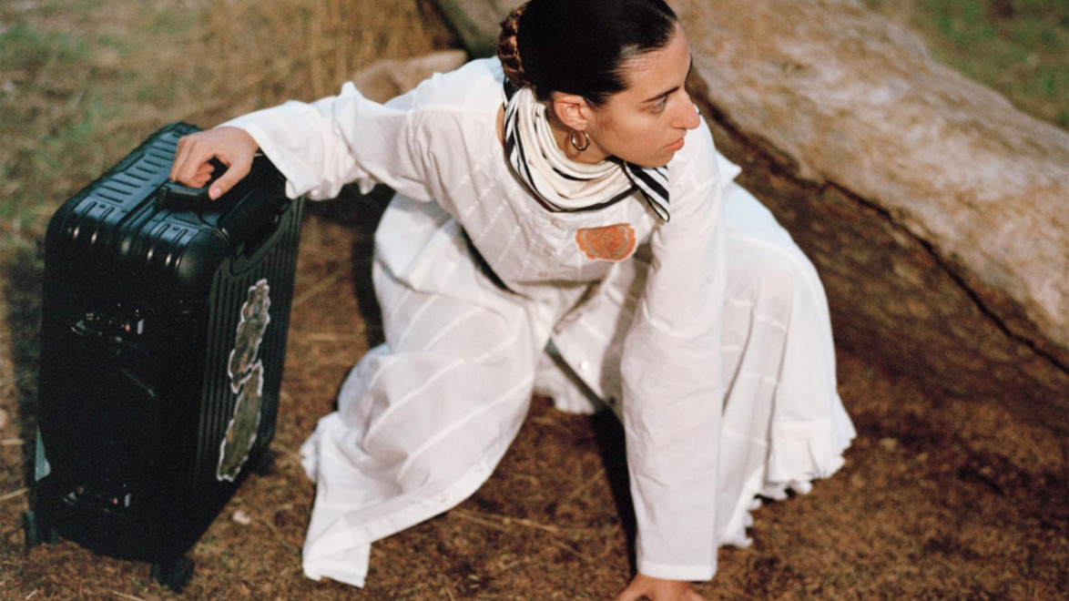 Woman in a white dress kneels on forest floor beside a black suitcase, with a large fallen tree in the background.