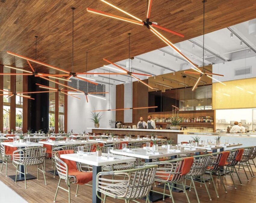 Modern restaurant interior with large wood ceiling, stylish angular light fixtures, white tables, and colorful chairs.