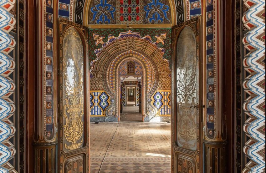 Ornate room with colorful, intricate patterns and geometric designs, seen through an open doorway in a historic building.