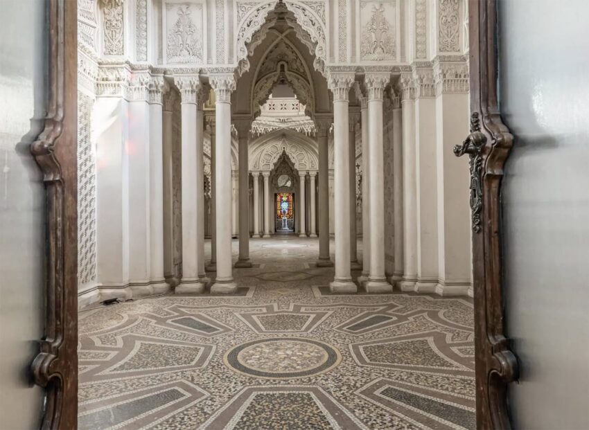 View of ornate hallway with intricate columns, detailed archways, and colorful stained glass window in the distance.