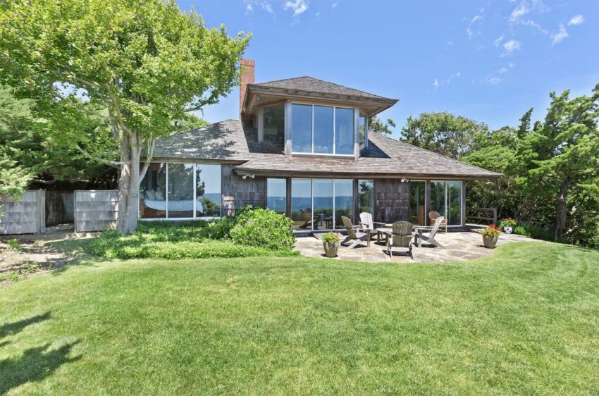 Modern house with large windows, surrounded by trees, features a stone patio with chairs on a sunny day.