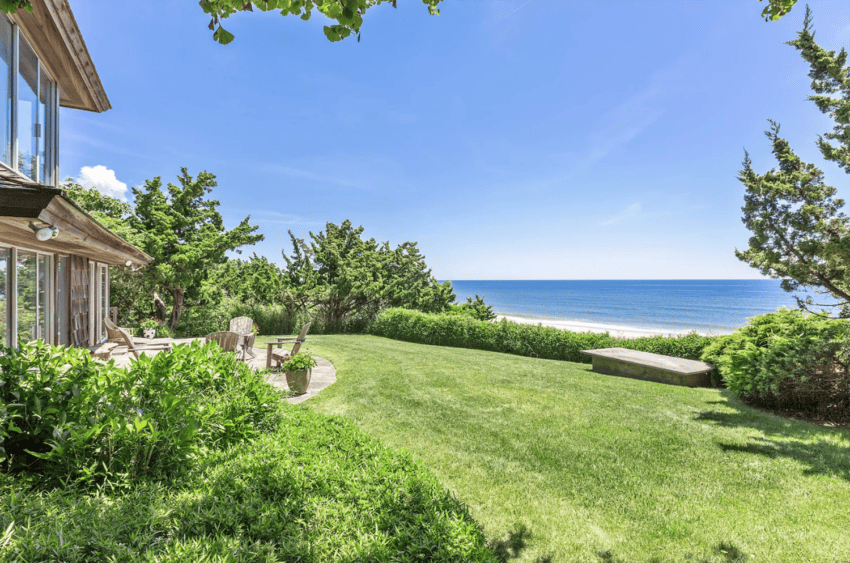 Coastal view with a grassy lawn, trees, and a house on the left overlooking the ocean on a sunny day.