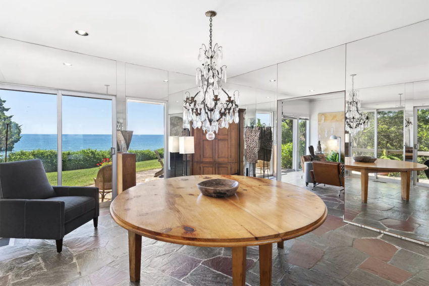 Elegant dining room with a round wooden table, chandelier, and mirrored walls overlooking a scenic ocean view.