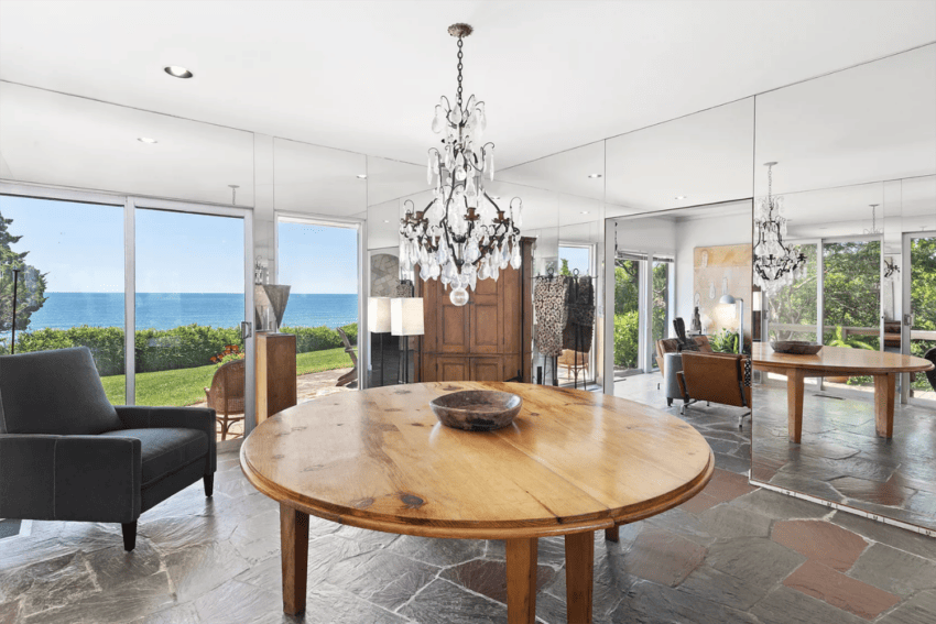 Elegant dining room with a round wooden table, chandelier, and mirrored walls overlooking a scenic ocean view.