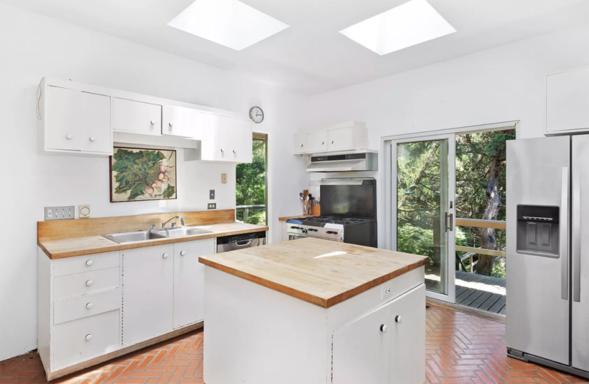 Bright white kitchen with a central island, wooden countertops, large fridge, and sliding doors leading to a balcony with greenery.
