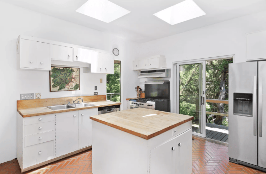Bright white kitchen with a central island, wooden countertops, large fridge, and sliding doors leading to a balcony with greenery.