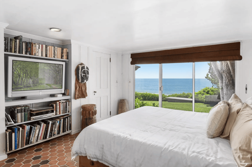 Cozy bedroom with sea view, features a bed, bookshelves, TV, and sliding door to a garden with ocean scenery.