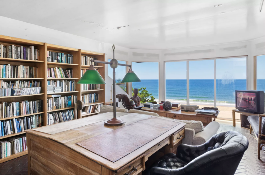 Cozy home library with bookshelves, wooden desk, leather chairs, and large windows offering a view of the ocean.