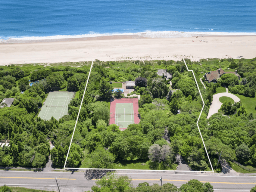 Aerial view of a beachfront property with tennis courts, green trees, and houses along a sandy beach.