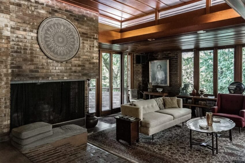 Cozy living room with brick walls, large fireplace, sofa, armchair, and large windows overlooking greenery.