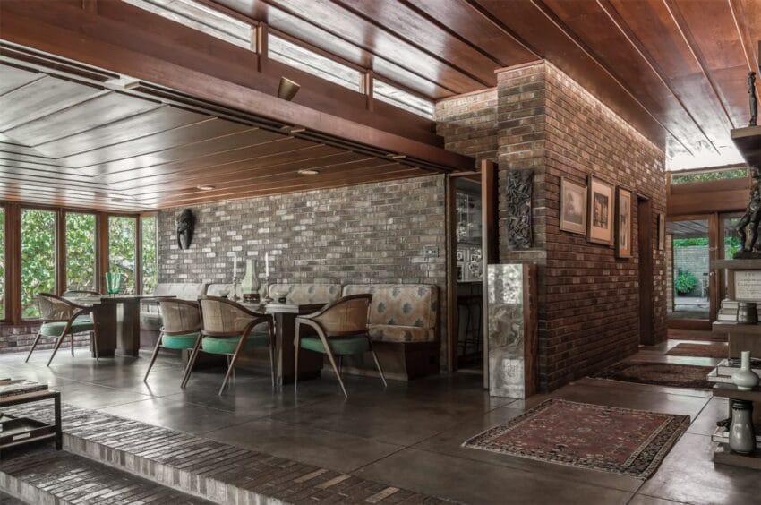 Mid-century modern dining room with brick walls, wooden ceiling, and wicker chairs around a table, with natural light.