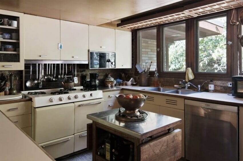 Vintage kitchen with cream appliances, large windows, and wooden accents. The counter holds a fruit bowl and kitchen utensils.