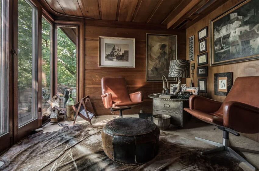 Cozy rustic wood-paneled room with leather chairs, framed art, large windows, zebra-patterned lamp, and cowhide rug.