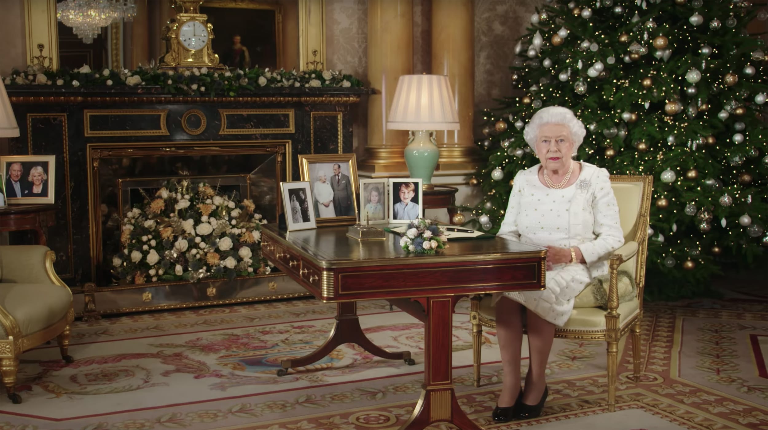 Elderly woman in white sitting at a desk with family photos, surrounded by Christmas decorations and a decorated tree.