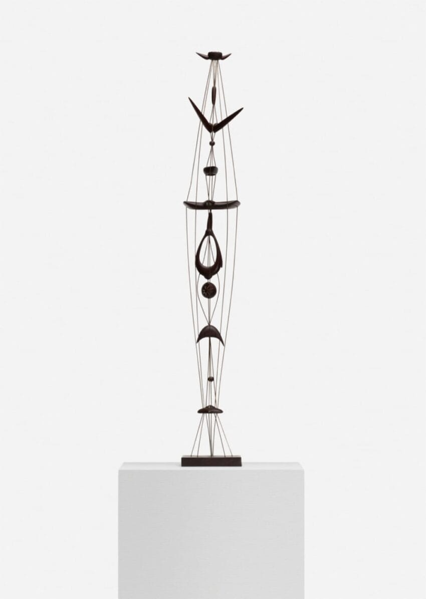 Tall abstract metal sculpture with geometric shapes on a white pedestal against a plain background.