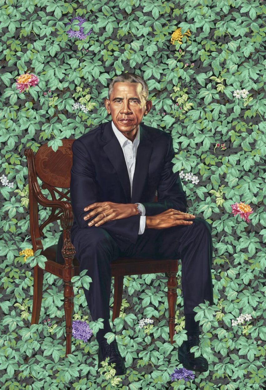 Man in black suit sitting on a wooden chair against a lush green leafy background with colorful flowers.