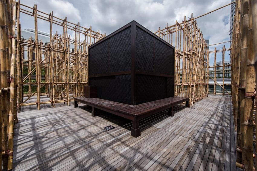 A modern art installation featuring a black cube surrounded by bamboo structures on a wooden deck under a cloudy sky.