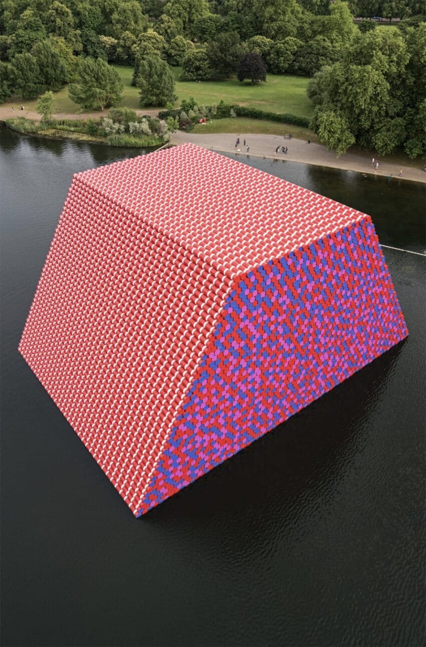 Large artistic installation made of colorful barrels floating on a serene lake, surrounded by green trees and a grassy area.