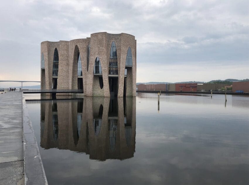 Modern building with curved arches reflected in calm water under a cloudy sky.