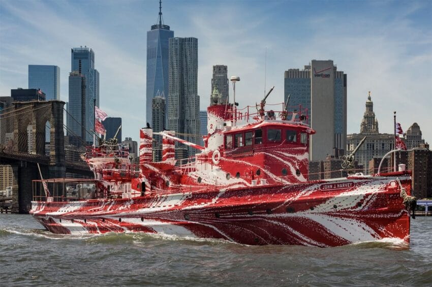 Red and white painted boat sailing on a river with a city skyline and bridge in the background.