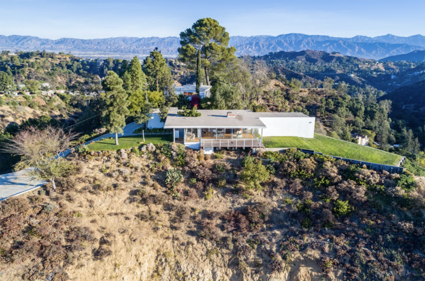 A modern house with a flat roof on a hill surrounded by trees, overlooking a mountainous landscape in daylight.