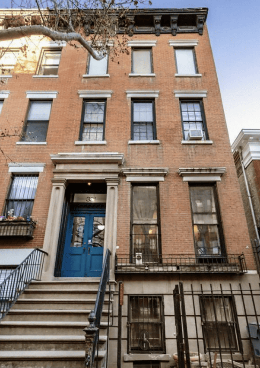 Four-story brownstone building with a blue door, black iron railings, and steps leading to the entrance. Tree branches overhead.