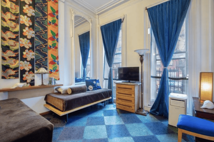 Colorful living room with blue curtains, patterned wall art, brown sofa, wooden furniture, and TV by large windows.