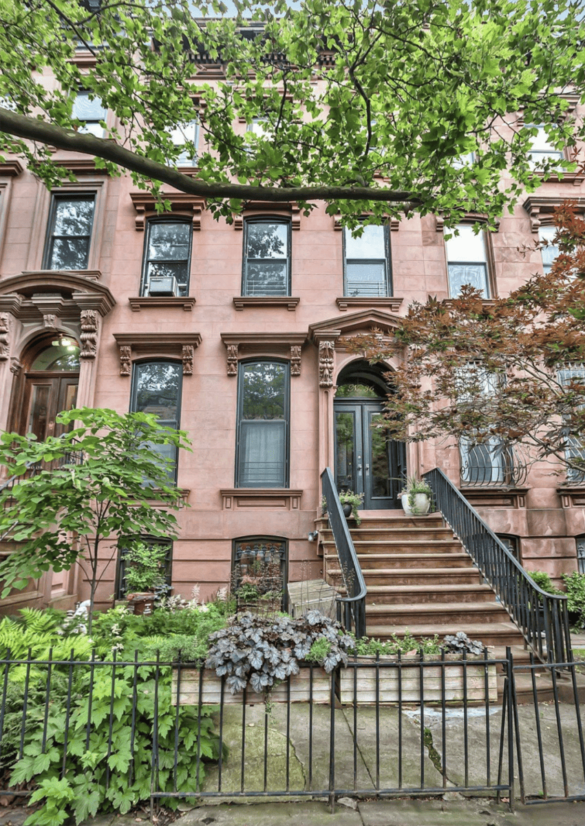 Exterior view of a classic brownstone building with ornate moldings, black railings, and lush greenery in the foreground