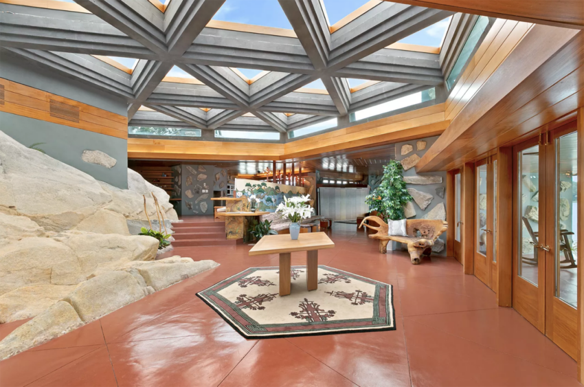 Interior of a modern home with geometric ceiling, large windows, stone walls, and a central table with a patterned rug.