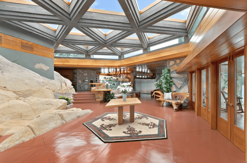 Interior of a modern home with geometric ceiling, large windows, stone walls, and a central table with a patterned rug.