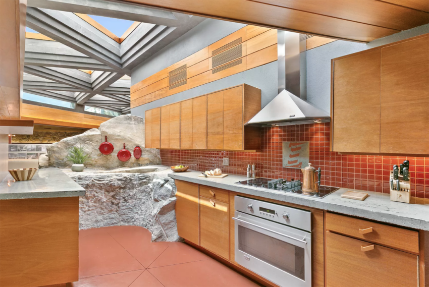 Modern kitchen with wooden cabinets, red tile backsplash, and natural rock integrated into the design under a skylight.