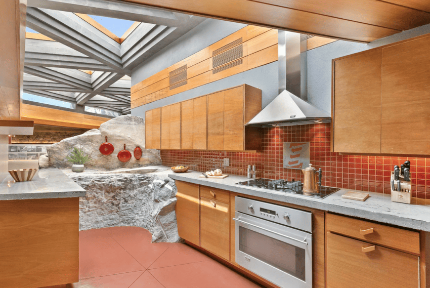 Modern kitchen with wooden cabinets, red tile backsplash, and natural rock integrated into the design under a skylight.