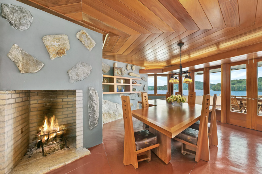 Wood-paneled dining room with stone fireplace, large table, and lake view through expansive windows. Warm and inviting atmosphere.