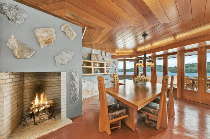 Wood-paneled dining room with stone fireplace, large table, and lake view through expansive windows. Warm and inviting atmosphere.