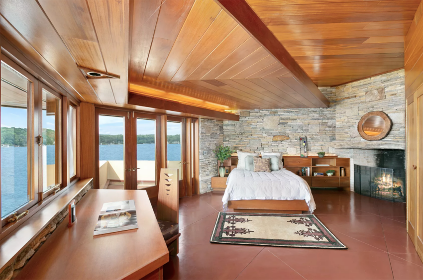 Wooden bedroom interior with large windows, stone fireplace, desk, and a view of the lake.