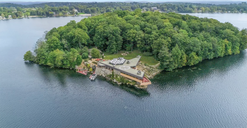 Isolated house surrounded by trees on a small island in a lake, with lush greenery and blue waters visible in the background.