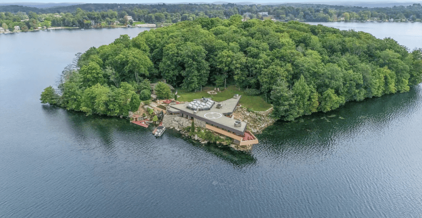 Isolated house surrounded by trees on a small island in a lake, with lush greenery and blue waters visible in the background.