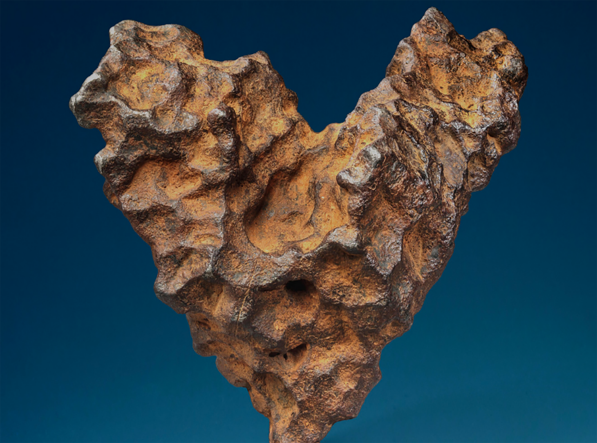 Rust-colored, heart-shaped meteorite with a rough texture against a dark blue background.