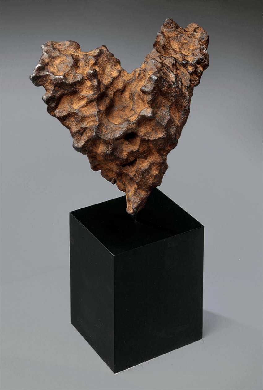 Heart-shaped meteorite displayed on a black pedestal against a gray background.