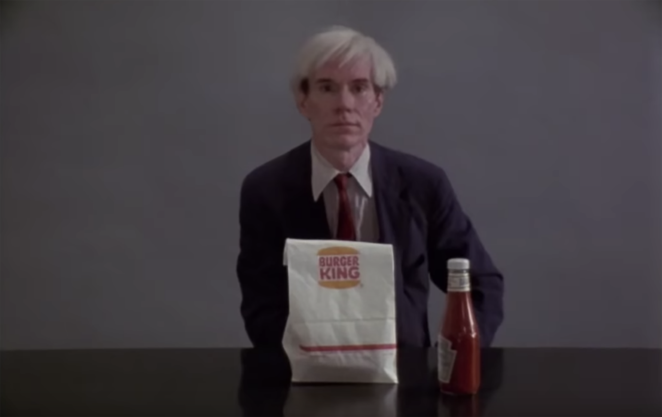 Man sitting at a table with a Burger King bag and a bottle of ketchup in front of a plain background.