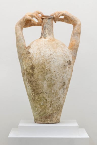 Sculpture of large clay vase with human-like hands grasping the top, displayed on a white pedestal against a plain background.